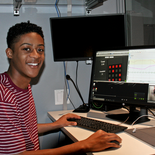 Photo of undergraduate student Marlon Washington II sitting in a dim observation room, with images from a video feed and corresponding data on his computer screen.