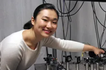 Smiling Asian female graduate student with her hair tied back, wearing a white sweater and leaning down over a complex microscope/laser contraption on a silver table.