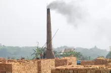Photo of a tall kiln tower streaming smoke with many huge stacks of orange/red bricks in front of it.