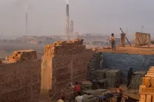 Photo showing workers moving bricks, surrounded by walls made of other bricks, with kilns smoking in the background.