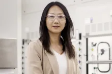 Photo of an Asian female faculty member standing in a very white-toned wet laboratory area, wearing clear protective safety goggles and a beige blazer.